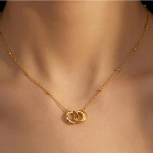 May include: A gold necklace with a pendant of two intertwined circles. The chain is fine, with tiny beads at intervals. The pendant has a small, clear gemstone at the centre of the interlocking circles.