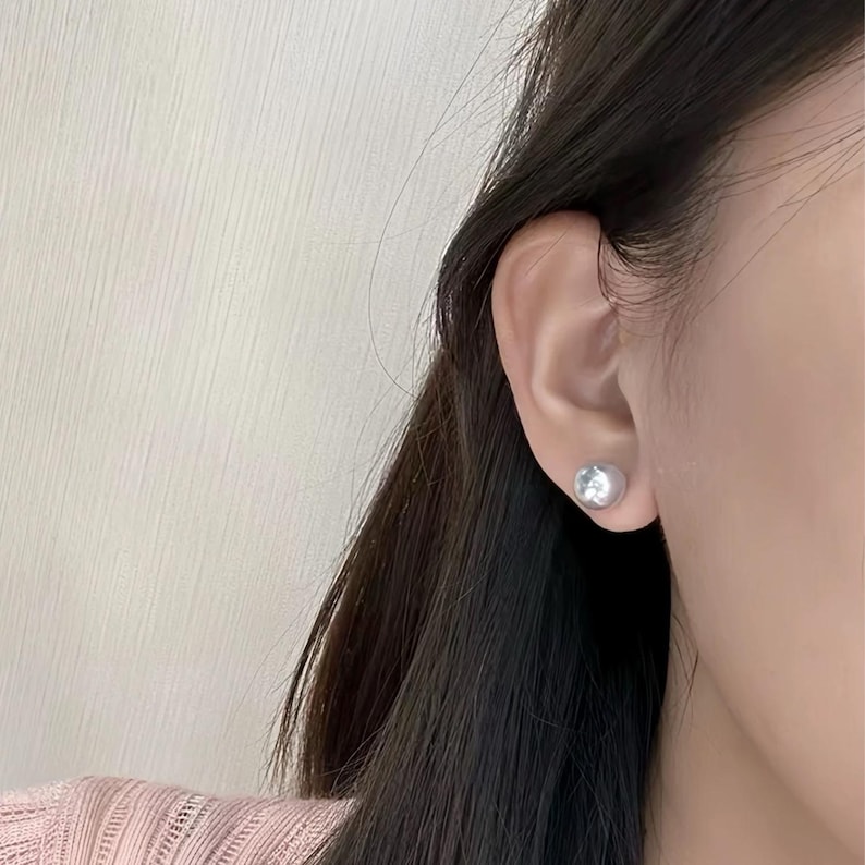 Puede incluir: Primer plano de un pendiente de bot&oacute;n plateado en una oreja. El pendiente es una peque&ntilde;a esfera redonda y brillante. La oreja est&aacute; situada en el lado derecho del marco, con pelo oscuro parcialmente visible a la izquierda.