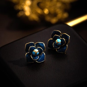May include: A pair of flower-shaped stud earrings. The earrings have a dark blue enamel finish with gold-coloured trim. Each flower's centre has a small, light blue stone. The earrings are displayed on a black surface.