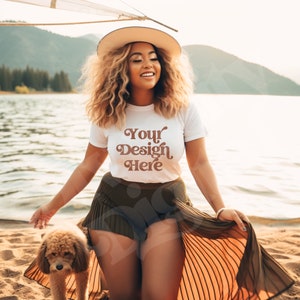 Op de afbeelding: Een vrouw draagt een wit T-shirt met de tekst "Your Design Here" in bruine letters, een strohoed en een bruine rok. Ze staat op een strand met een kleine hond.