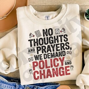 以下が含まれることがあります： 「NO THOUGHTS AND PRAYERS, WE DEMAND POLICY AND CHANGE」のテキストが黒と赤でプリントされたクリーム色のスウェットシャツ。メガホン、チェックリスト、拳などのグラフィック要素が含まれています。