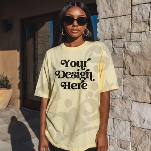 May include: A light yellow t-shirt with the text "Your Design Here" in a black, decorative font. The shirt is short-sleeved and appears to be made of a soft material. The model is wearing sunglasses and gold hoop earrings.
