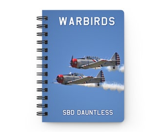 May include: A blue notebook with a spiral binding features a photo of two vintage airplanes flying in formation against a blue sky. The text "WARBIRDS" is at the top of the notebook and "SBD DAUNTLESS" is at the bottom.