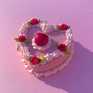 May include: A pink heart-shaped cake with white frosting and red roses. The cake has a red cherry on top.