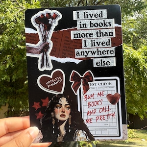 Puede incluir: Una tarjeta decorativa negra con varias pegatinas y texto. La tarjeta presenta un ramo de rosas rojas, una pegatina en forma de coraz&oacute;n con las palabras "HOPELESS ROMANTIC" y un dibujo de una mujer. El texto en la tarjeta dice "I lived in books more than I lived anywhere else."