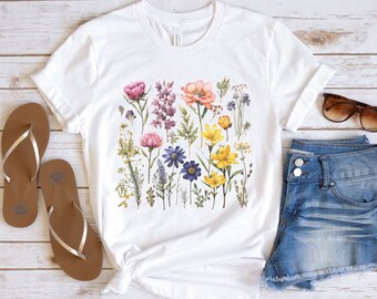 Op de afbeelding: Wit T-shirt met een aquarel bloemenprint in roze, paars, geel en blauw. Het shirt wordt gecombineerd met een spijkerbroekshort, bruine sandalen en een zonnebril, wat een casual, zomerse look creëert.