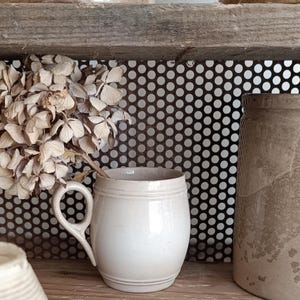 Peut inclure: Une tasse en céramique crème avec une anse incurvée et des rainures horizontales. La tasse est posée sur une étagère en bois avec des fleurs séchées et un fond en métal perforé. D'autres articles en céramique sont également visibles.