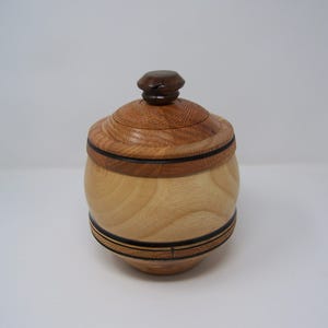 May include: A handcrafted wooden jar with a lid. The jar is made of light and dark wood, with a round body and a decorative band. The lid has a small, dark brown knob. The jar is likely used for storage.