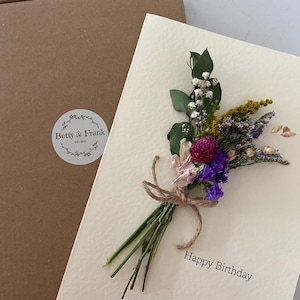 May include: A cream-coloured birthday card adorned with a dried flower bouquet tied with twine. The card reads "Happy Birthday" in black. A brown box with a "Betty & Frank" sticker is visible in the background.