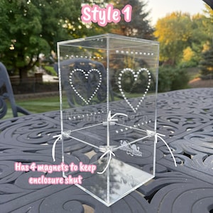 May include: Clear acrylic box with heart-shaped dot designs. The box has the text "Style 1" and "Has 4 magnets to keep enclosure shut". The box is on a decorative black table.