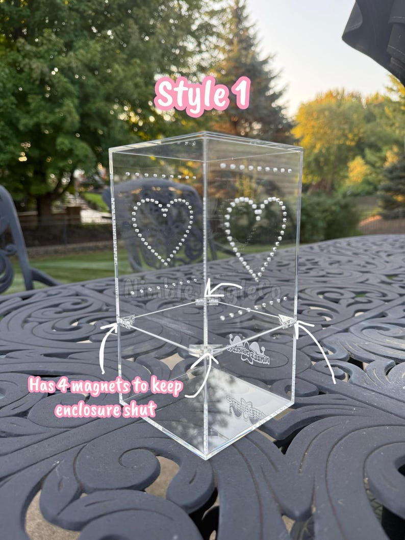 May include: Clear acrylic box with heart-shaped dot designs. The box has the text "Style 1" and "Has 4 magnets to keep enclosure shut". The box is on a decorative black table.
