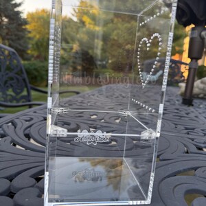 May include: A clear acrylic box with a heart design made of small dots. The box has the text "NimbleJingleCo" on the side and bottom. The box is on a dark, patterned surface.