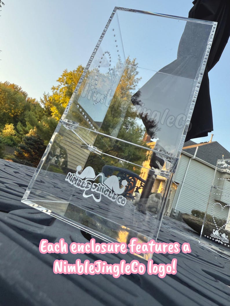 May include: Clear acrylic display case with a NimbleJingleCo logo. The case is angled, showing the interior and exterior details. The text "Each enclosure features a NimbleJingleCo logo!" is displayed in pink.
