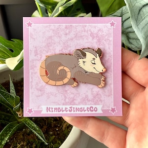 May include: A pink and white card with a gold star design features a gold-colored enamel pin of a brown and white opossum curled up sleeping. The pin is on a white background and the card has the text "Nimble Jingle Co."