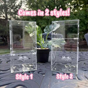 May include: Two clear acrylic display cases, labeled Style 1 and Style 2, are shown side-by-side. The cases have a rectangular shape and are designed to showcase items. A small potted plant sits between them. The text "Comes in 2 styles!" is above the cases.