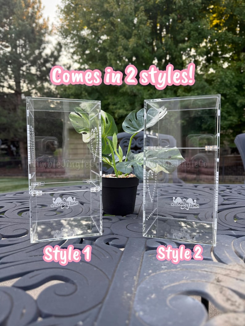 May include: Two clear acrylic display cases, labeled Style 1 and Style 2, are shown side-by-side. The cases have a rectangular shape and are designed to showcase items. A small potted plant sits between them. The text "Comes in 2 styles!" is above the cases.