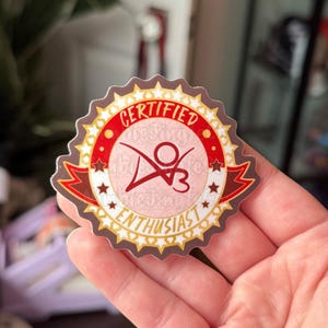 May include: A round sticker with a red and gold design. The sticker reads "CERTIFIED ENTHUSIAST" in a serif font. The sticker has a light pink center with a stylized graphic and the letters "d3".