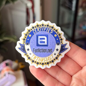 May include: A white sticker with a blue and gold design. The sticker has the words "CERTIFIED" and "ENTHUSIAST" in a circular pattern. The center features the text "Fanfiction.net" and a stylized letter "E".