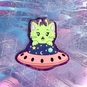May include: A green alien cat with antennas sits in a pink flying saucer with a blue and purple starry background.