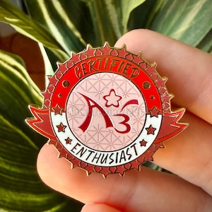 Op de afbeelding: Een rode en gouden emaille pin met de woorden "CERTIFIED" en "ENTHUSIAST" rond een centraal ontwerp. Het ontwerp bevat de letters "A3" en een ster. De pin wordt in een hand gehouden.