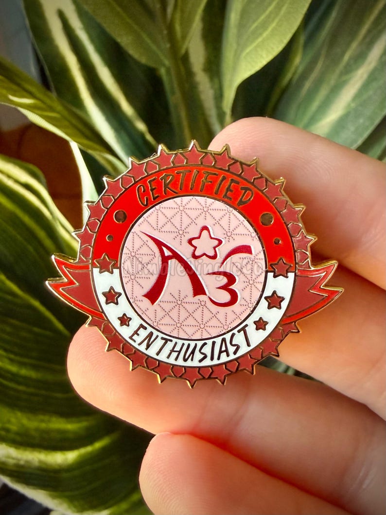 Op de afbeelding: Een rode en gouden emaille pin met de woorden "CERTIFIED" en "ENTHUSIAST" rond een centraal ontwerp. Het ontwerp bevat de letters "A3" en een ster. De pin wordt in een hand gehouden.