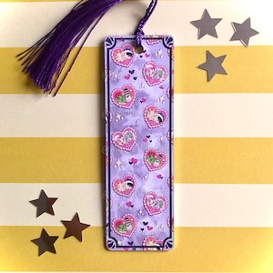Puede incluir: Un marcapáginas rectangular con una borla morada. El marcapáginas presenta un patrón repetido de ilustraciones en forma de corazón en rosa, verde y morado. El fondo es de color lila claro con pequeñas estrellas.
