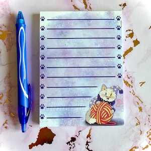 Op de afbeelding: Een paars-wit notitieboekje met een kattenillustratie. De kat draagt een hoed en houdt een bol wol vast. Het notitieboekje heeft een gestreept ontwerp en poot afdruk randen.