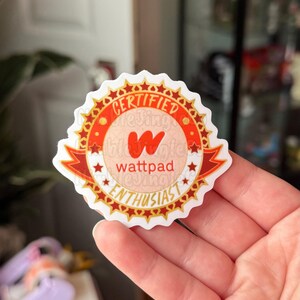 May include: A round sticker with a red and gold design. The sticker reads "CERTIFIED" at the top, "wattpad" in the center, and "ENTHUSIAST" at the bottom. The sticker has a white border and is held by a hand.