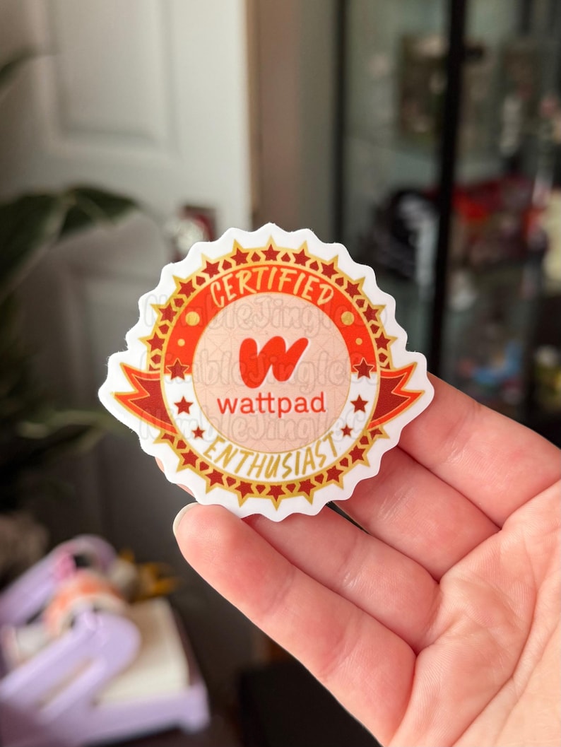 May include: A round sticker with a red and gold design. The sticker reads "CERTIFIED" at the top, "wattpad" in the center, and "ENTHUSIAST" at the bottom. The sticker has a white border and is held by a hand.