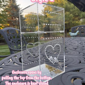 May include: Clear acrylic jewelry display case, labeled "Style 2", with heart-shaped stud earring holders. The enclosure opens by pulling the top from the bottom and is kept closed by 4 magnets. The display case is on a decorative table.