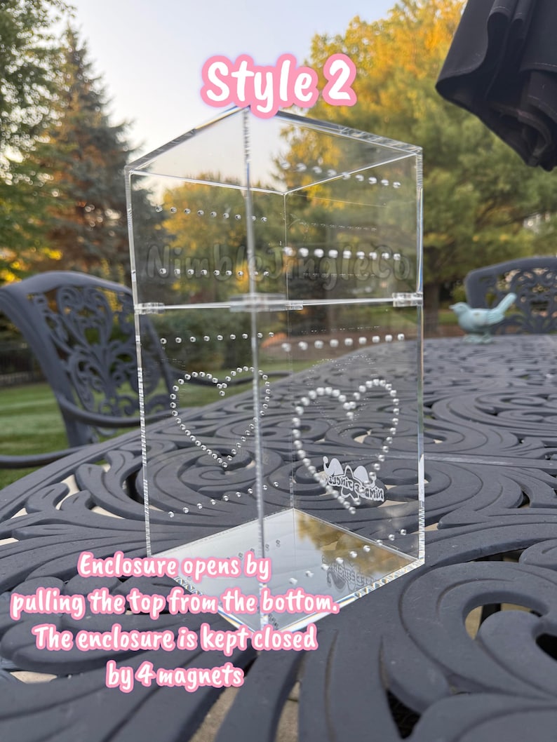 May include: Clear acrylic jewelry display case, labeled "Style 2", with heart-shaped stud earring holders. The enclosure opens by pulling the top from the bottom and is kept closed by 4 magnets. The display case is on a decorative table.
