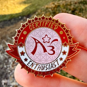 Op de afbeelding: Rode en gouden emaille pin met de tekst "CERTIFIED ENTHUSIAST" en de letters "A3". De pin heeft een sterrenrand en een lintbanner-ontwerp, met een roze en witte achtergrond.