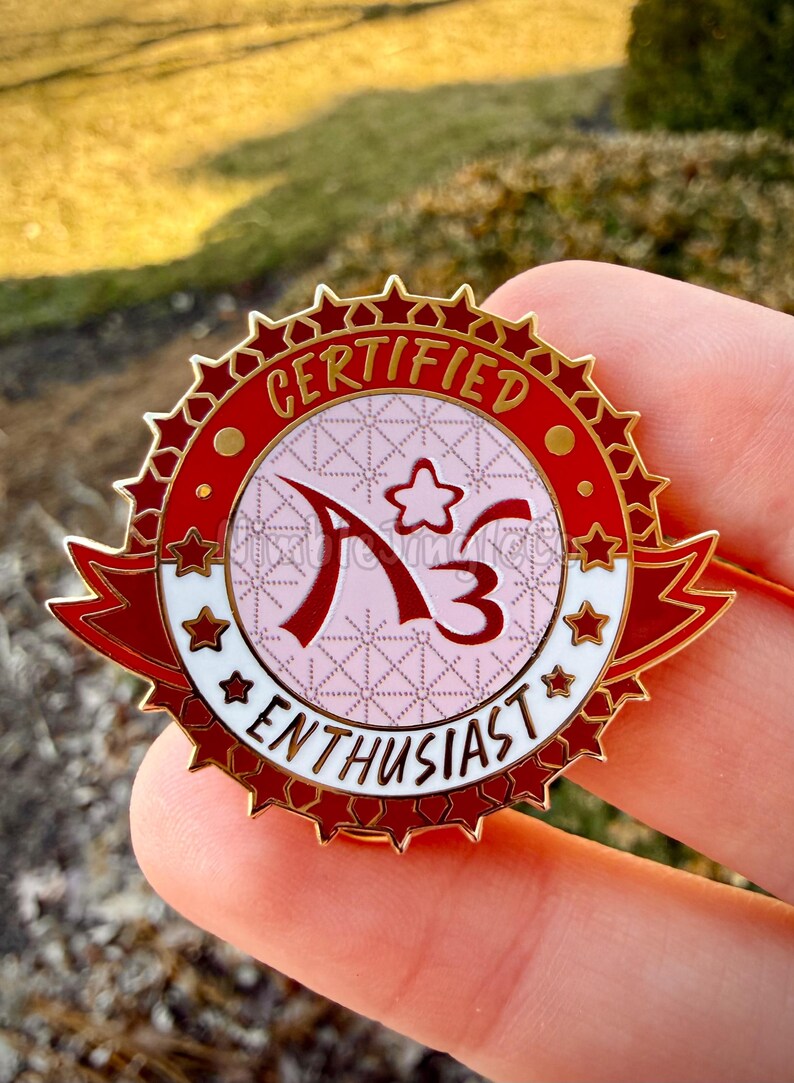 Op de afbeelding: Rode en gouden emaille pin met de tekst "CERTIFIED ENTHUSIAST" en de letters "A3". De pin heeft een sterrenrand en een lintbanner-ontwerp, met een roze en witte achtergrond.