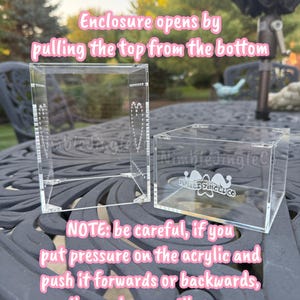 May include: Two clear acrylic display boxes are shown. The larger box is open, and the smaller box is closed. Text on the image reads: "Enclosure opens by pulling the top from the bottom." and "NOTE: be careful, if you put pressure on the acrylic and push it forwards or backwards, the enclosure will open."