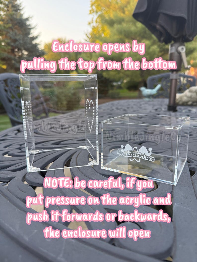 May include: Two clear acrylic display boxes are shown. The larger box is open, and the smaller box is closed. Text on the image reads: "Enclosure opens by pulling the top from the bottom." and "NOTE: be careful, if you put pressure on the acrylic and push it forwards or backwards, the enclosure will open."