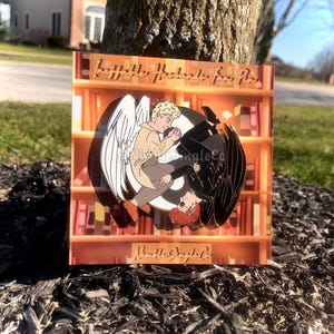 May include: A gold enamel pin featuring two figures, one with angel wings and the other with devil wings, embracing. The pin is set against a background of a bookshelf with the text "Love Hate Husbands Fan Pin" and "Nible Jingle Co."