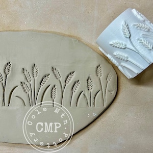 May include: A white ceramic rolling pin with a wheat design is shown next to a clay slab with the same design impressed into it. The clay slab has the text "Oole Mtn. Pottery CMP" stamped on it.