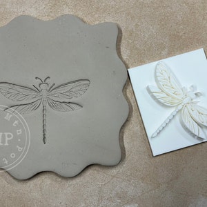 May include: A gray clay mold with a dragonfly design and the text "Cool Mountain Pottery CMP" on the bottom. Next to the mold is a white silicone mold with a dragonfly design.