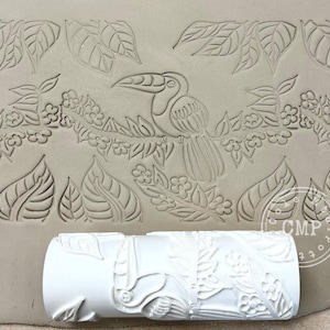 May include: A white ceramic roller with a toucan and floral design, used to create an embossed pattern on clay. The roller is shown next to a clay slab with the embossed toucan and leaf design.