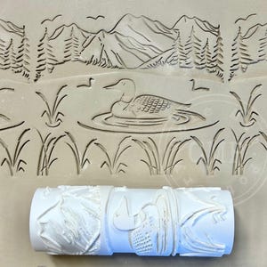 May include: A white roller with a nature-themed design. The design includes a lake scene with mountains, trees, birds, and a loon. The roller is used for creating patterns on surfaces. The overall theme is nature and outdoor scenes.