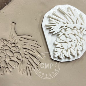 May include: A white ceramic pine cone stamp with a detailed design. The stamp is used to create an impression of a pine cone in clay.