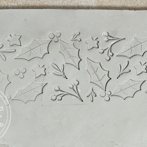 May include: A green rolling pin with a holly leaf and berry design is used to create an imprint of the design on a light gray clay surface.