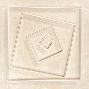 May include: A set of four white square frames, each nested inside the other, creating a geometric pattern. The frames are arranged in a diagonal pattern, with the smallest frame in the center.
