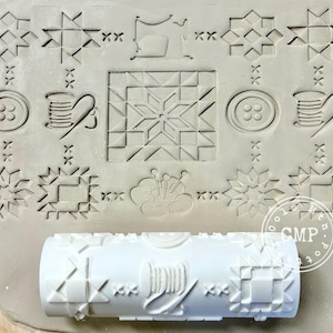 May include: A white rolling pin with a geometric pattern and a spool of thread design is shown on a clay surface with various embossed sewing-themed designs. The designs include stars, buttons, and sewing notions.
