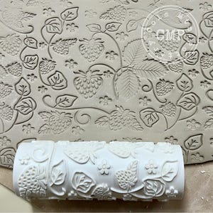 May include: A white rolling pin with a raised design of strawberries and leaves. The rolling pin is being used to create an impression in a piece of clay.