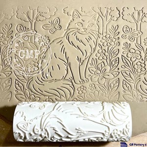 May include: A beige clay slab with an embossed design of a cat, flowers, trees, and butterflies. A white roller with a floral pattern is in front of the slab. The slab has the letters "CMF" in a circle.
