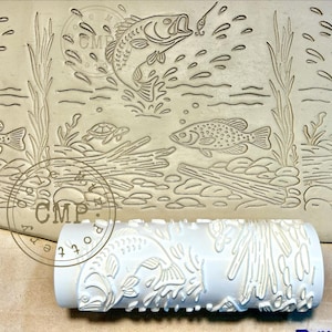 May include: A white cylindrical clay texture roller featuring a raised relief pattern of a jumping fish, a swimming fish, a small turtle, aquatic plants, and logs. The design is imprinted into a flat clay surface, showing the detailed underwater scene.