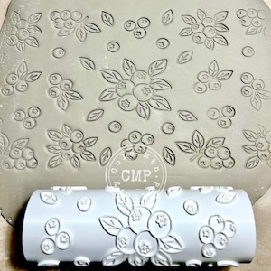 May include: A white rolling pin with a blueberry design is shown next to a clay slab with the same embossed pattern. The rolling pin has a cylindrical shape with a repeating pattern of blueberries and leaves. The clay slab has a similar pattern.