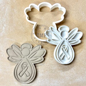 May include: Two angel-shaped cookie cutters, one white and one beige, with a teardrop-shaped body and a ribbon design. The white cutter is next to a cloud-shaped cutter. The beige cutter is a finished cookie.