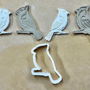 May include: Five white ceramic cardinal bird cookie cutters, including one larger cutter in the center. The cutters are on a brown surface.